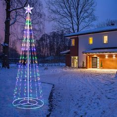 a lighted christmas tree in front of a house with snow on the ground and trees around it