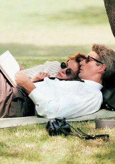 a man laying on the ground next to a tree while holding a baby and reading a book