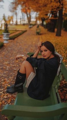 a woman sitting on top of a green bench next to trees with leaves all over the ground