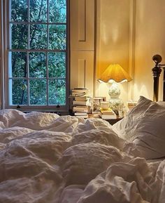 an unmade bed in front of a window with a lamp on the end table