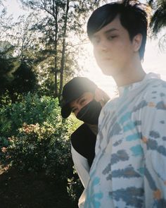 two young men standing next to each other near trees and bushes with the sun shining on them