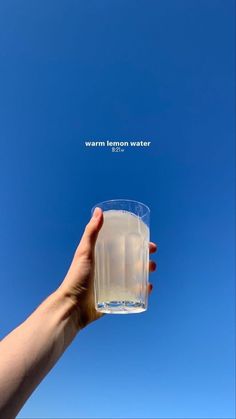 a hand holding up a glass with water in it against a blue sky and the words warm lemon water