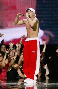 a shirtless man in red and white pants singing into a microphone with his hands up