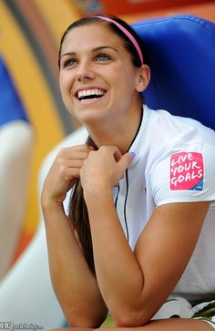 a smiling woman wearing a white shirt and pink headband with live your goals sticker on it