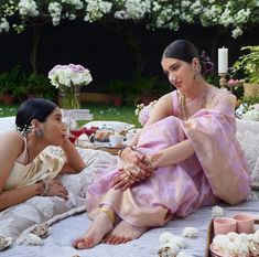 two women sitting on a bed with flowers in the background and food spread out around them