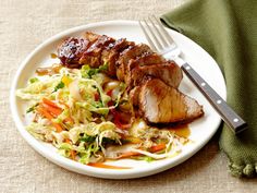 a white plate topped with meat and coleslaw next to a knife and fork