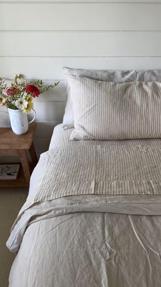 a bed with white sheets and pillows next to a wooden side table filled with flowers