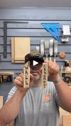 a man holding up two pieces of wood in front of a wall with tools on it