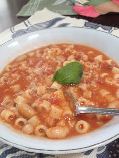 a white bowl filled with pasta and sauce on top of a blue table cloth next to a silver spoon