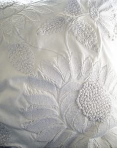 an embroidered pillow with white flowers and leaves on the front, sitting on a bed