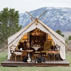 two dogs are sitting in front of a teepee tent with mountains in the background