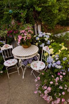 an outdoor table and chairs are surrounded by flowers in the garden, including hydrangeas