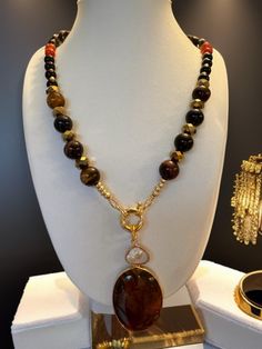 The necklace in the image showcases a striking central amber-colored oval pendant, suspended from a gold-tone ring that also holds a smaller, translucent pearl, creating a dual charm effect. The beads, in varying shades from deep browns to warm amber and occasional hints of red, are faceted for a gentle sparkle. The assembly is completed with a gold-tone beads for a touch of elegance followed  by tiger eye  12 mm grade A beads and 8mm of black obsidian , gold faceted rondelles, red agate represe Red Agate, Black Obsidian, Amber Color, Oval Pendant, Pendant Design, Love Necklace, Jewelry Lover, Tiger Eye, Limited Time