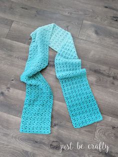 a blue crocheted scarf laying on top of a wooden floor with the words just be crafty written across it