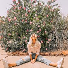 a woman sitting on the ground in front of a bush with pink flowers behind her