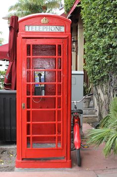 a red phone booth sitting on the side of a road next to a bike and trees