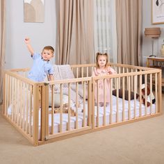 two children standing in a wooden crib with curtains on the windowsill and one child holding his hand up