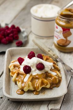 a waffle topped with whipped cream and raspberries on a plate next to a jar of peanut butter