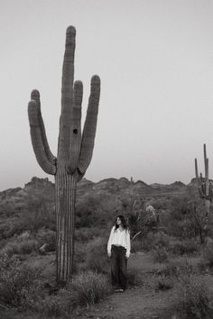 a person standing next to a large cactus in the middle of a dirt field with other plants