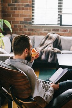 a man sitting in a chair reading a book and drinking from a cup while looking at his laptop