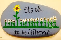 a painted rock that says it's ok to be different with daisies in the foreground