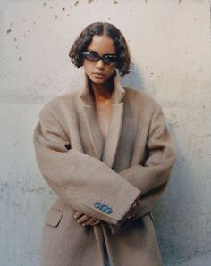 a woman with her arms crossed standing in front of a wall wearing sunglasses and a coat