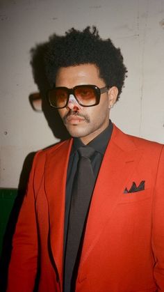 a man in a red suit and black tie with sunglasses on his face, standing next to a wall