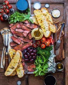 an assortment of meats, bread and vegetables on a wooden table with utensils