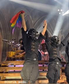 three men in black shirts and helmets on stage with their hands up to the sky