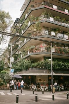 people sitting at tables in front of a tall building with plants growing on the balconies