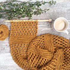 a crocheted scarf next to a cup of coffee on a wooden table with greenery