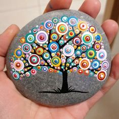 a hand holding a painted rock with a tree on it's face and colorful circles around the tree