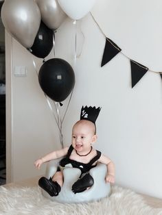 a baby sitting on top of a white chair with balloons in the air behind it