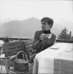 a woman sitting on a bench holding a cup and looking at the camera with mountains in the background