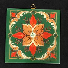 a green and orange square tile with flowers on the bottom is hanging from a hook