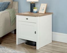 a white nightstand with a wooden top in a living room next to a couch and lamp