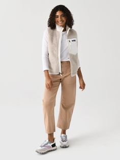 a woman standing in front of a white background wearing a beige vest and tan pants