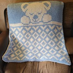 a blue and white knitted teddy bear pillow sitting on a brown couch next to a remote control