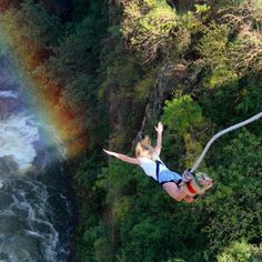 a woman flying through the air while riding a zip line over a river with a rainbow in the background