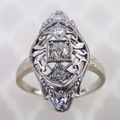 An old European cut diamond is centered on this antique 14k white gold filigree ring. The 3mm .10 carat precious gem is mounted in a square setting. Delicate airy scroll filigree with leaves and millgrain dots decorates the ring. The 13/16 inch long face has a domed gallery with engraved profiles, which is emulated on the ring's shoulders. There are four additional 1.5mm to 1.8mm diamonds set around on the ring. Collectively, this ring has an estimated total weight of .18 cwt diamonds.  The insi Victorian Diamond Ring With Rose Cut In White Gold, Victorian White Gold Diamond Ring With Rose Cut Diamonds, Victorian White Gold Diamond Ring With Rose Cut, Ornate Brilliant Cut Diamond Ring, Vintage Diamond Ring With Rose Cut Diamonds For Anniversary, Vintage Rose Cut Diamond Ring For Anniversary, Victorian Style White Diamond Ring, Vintage Diamond White Brilliant Cut Filigree Ring, Vintage Filigree Ring With Brilliant Cut In Diamond White