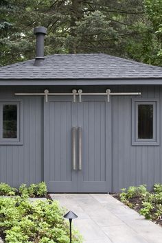 a small gray shed with two windows and a door on the side, surrounded by trees