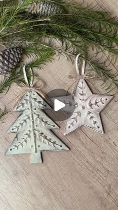 two metal christmas tree ornaments hanging on a wooden table next to pine cones and evergreen branches