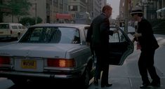 two men are talking to each other on the sidewalk next to a car and buildings