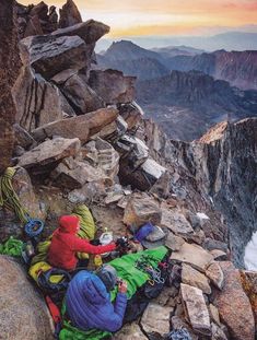 two people sitting on the side of a mountain with backpacks and sleeping bags in front of them