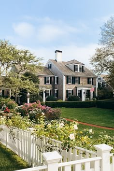 a white picket fence in front of a large house with lots of trees and flowers