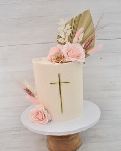 a white cake with pink flowers and a gold cross on the top is sitting on a stand