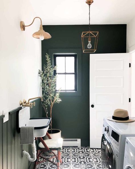 This modern laundry room displays dark wood wainscoting and a feature wall that contrasts with light white painted walls. Gold accents are found in the faucet above a vintage sink, as well as the light fixtures that illuminate the gray and white patterned tile floor. Laundry Room Paint Color, Laundry Room Paint, Wood Wainscoting, Wall Mount Kitchen Faucet, Vintage Sink, Green Laundry, Modern Laundry, Dark Green Walls, Patterned Tile