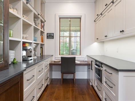 Custom-built by Morgan-Keefe Builders, this multi-functional space is a butler's pantry with a work desk. It offers a perfect place to write-up a grocery list and serves as a hidden drop-zone for that nonstop flow of mail that enters the home. Gray countertops contrast nicely with the bright white custom built-in shelves and exquisite cabinetry; while the natural wood built-in desk overlooks views of the exterior. Get more ideas for unique home features on our website. Working Butlers Pantry, Work Desk In Kitchen, Office In Pantry, Butler Pantry Office Combo, Butler Pantry With Window, Pantry Desk Combo, Pantry And Office Combo, Butlers Pantry Office Combo, Desk Nook In Kitchen