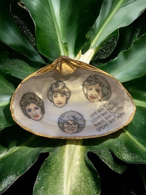 Celebrate your love for the iconic Golden Girls with this handcrafted decoupaged clam shell trinket dish featuring the beloved characters Rose, Blanche, Dorothy, and Sophia, along with the fun quote: "Live like Rose, Love like Blanche, Think like Dorothy, Speak like Sophia." Each natural clam shell is unique in shape and size, making this a one-of-a-kind piece. The hand-painted gold trim adds an elegant touch, making it perfect as a jewelry holder, desk accessory, or nostalgic decor piece. *🌟 Golden Girls Design: Features the iconic characters and their timeless charm, making it the perfect tribute for fans. *✨ Handcrafted & Unique: Each natural clam shell is individually decoupaged and gold-trimmed for a one-of-a-kind item. *💛 Versatile Use: Perfect for holding jewelry, small trinkets,