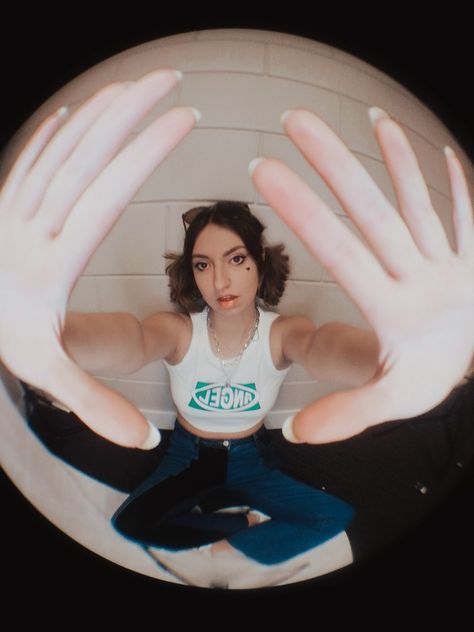 A girl is sitting in this picture with a white croptop and blue jeans. She has her short hair in two ponytails and has sunglasses up her head. She's holding her hands up close to the camera, which looks cool because there is a fish lens used. This picture has got 90s and 00s vibes to it. Funky Perspective Poses, Fish Eye Lense Drawings, Fishbowl Lens Aesthetic, Fish Lens Reference, Fisheye Perspective Drawing Face, Fisheye Art Tutorial, Fisheye Photoshoot Aesthetic, Fish Eye Lense Aesthetic, Fish Eye Poses Reference